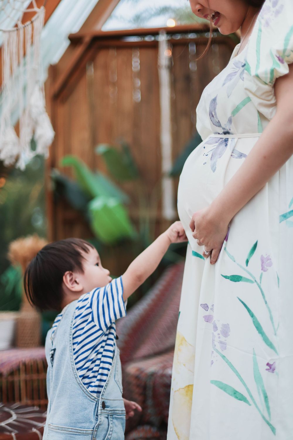 Pregnant woman in floral dress holds hands with toddler in striped shirt and denim overalls in a boho setting.