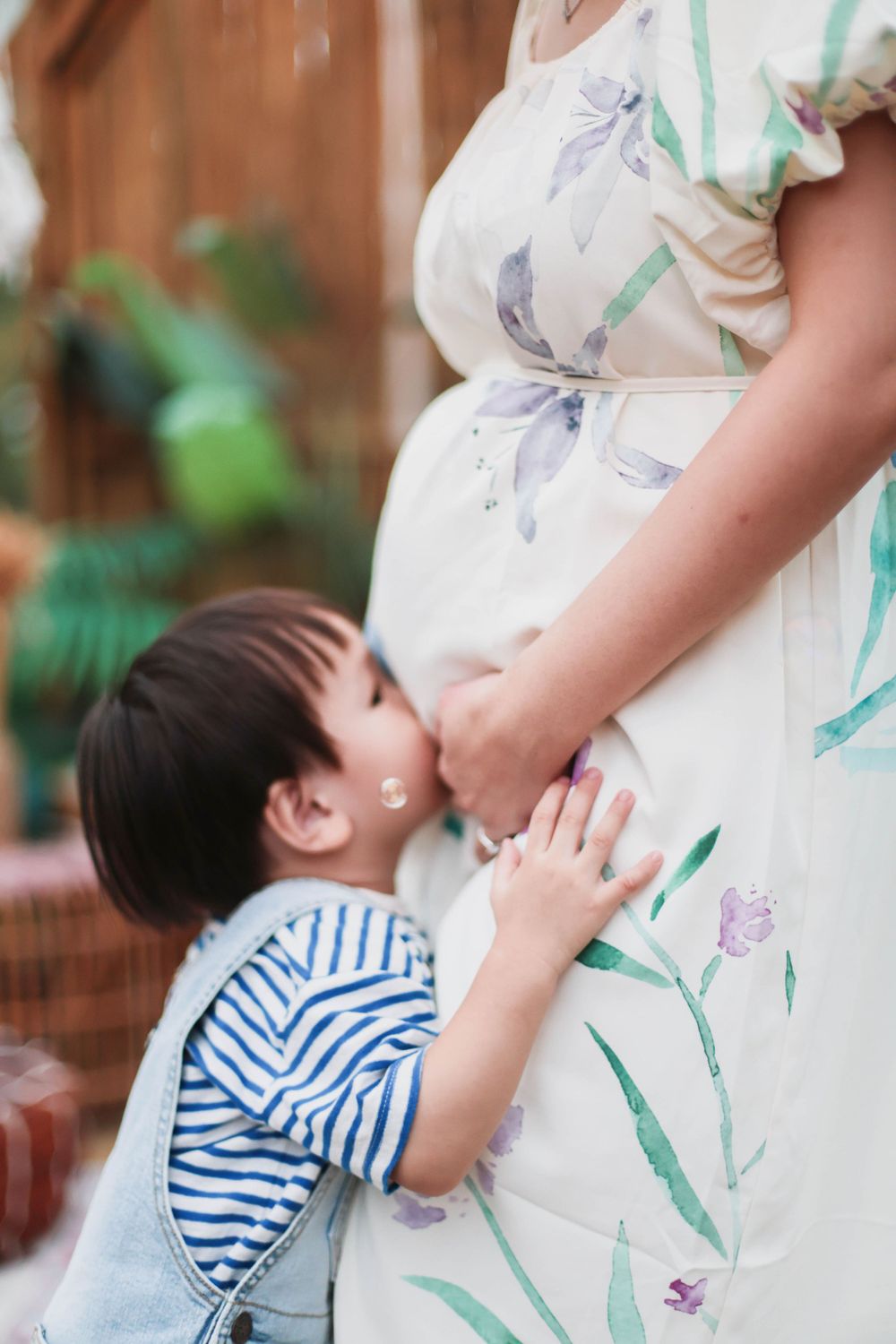 Young toddler boy kissing pregnant mothers belly in a beautiful floral maternity photo session.
