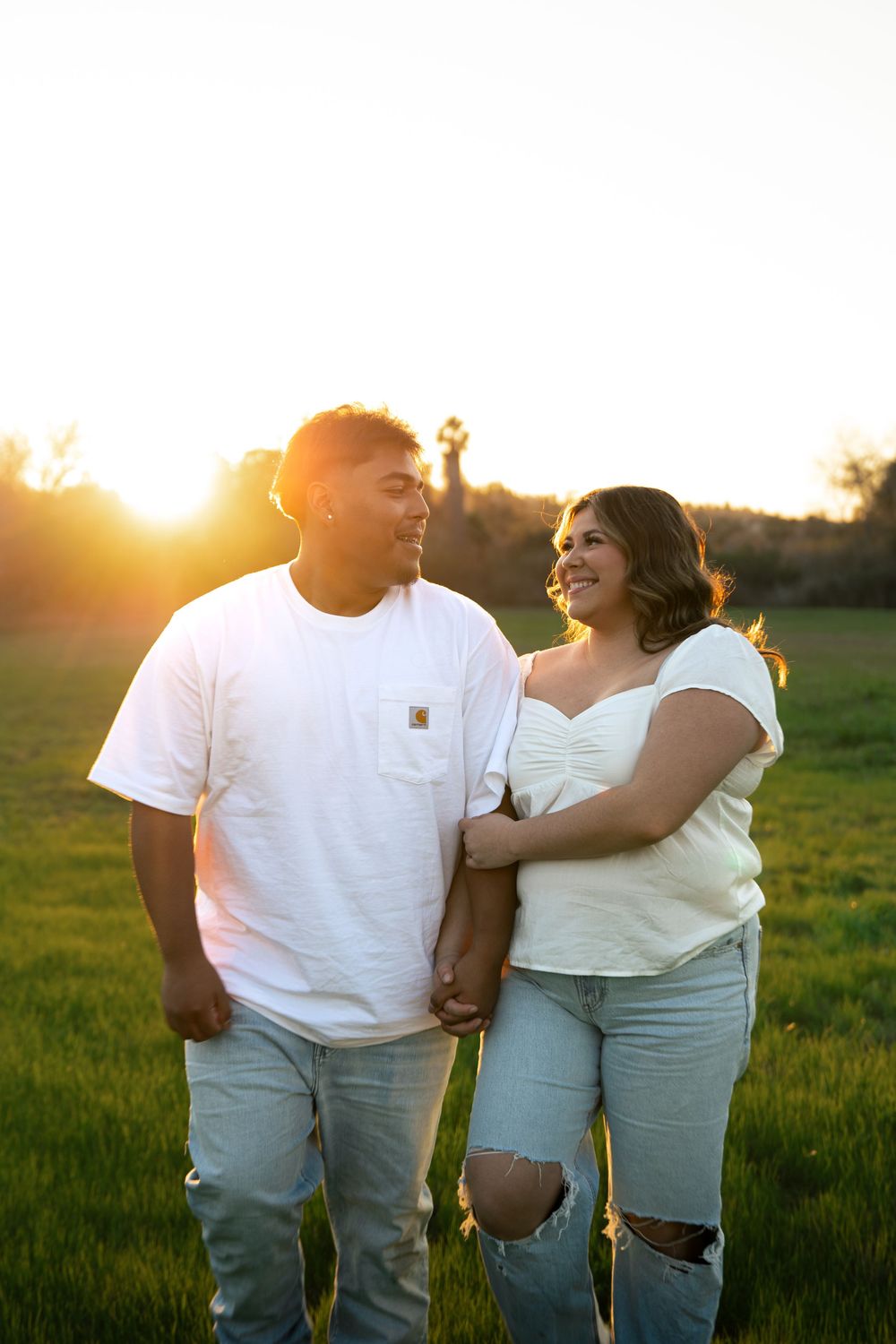 A couple in white outfits walks through a grassy field at sunset, creating a romantic silhouette against the golden sky.