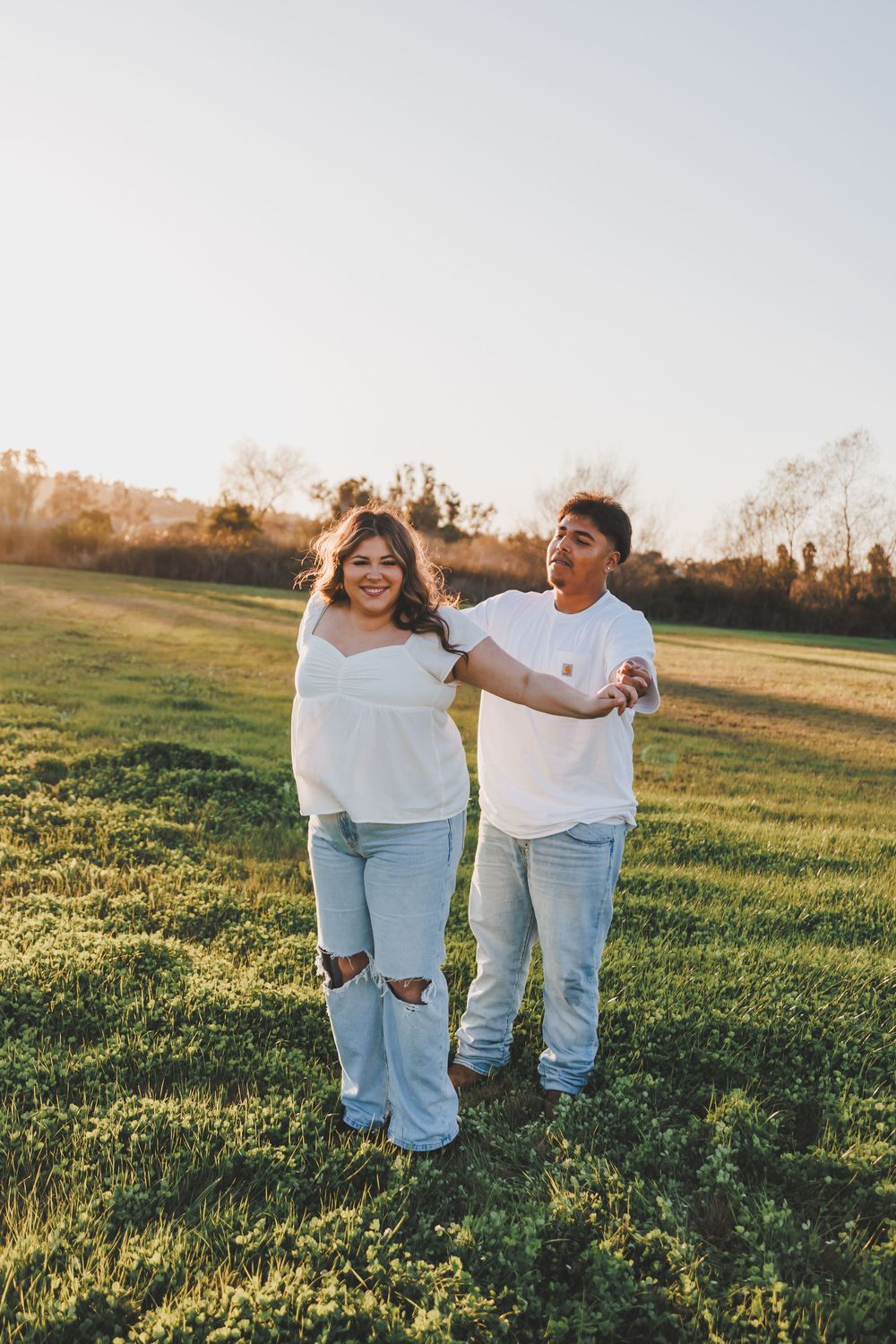 Two people in white shirts and jeans walk together through a sunlit meadow at golden hour.