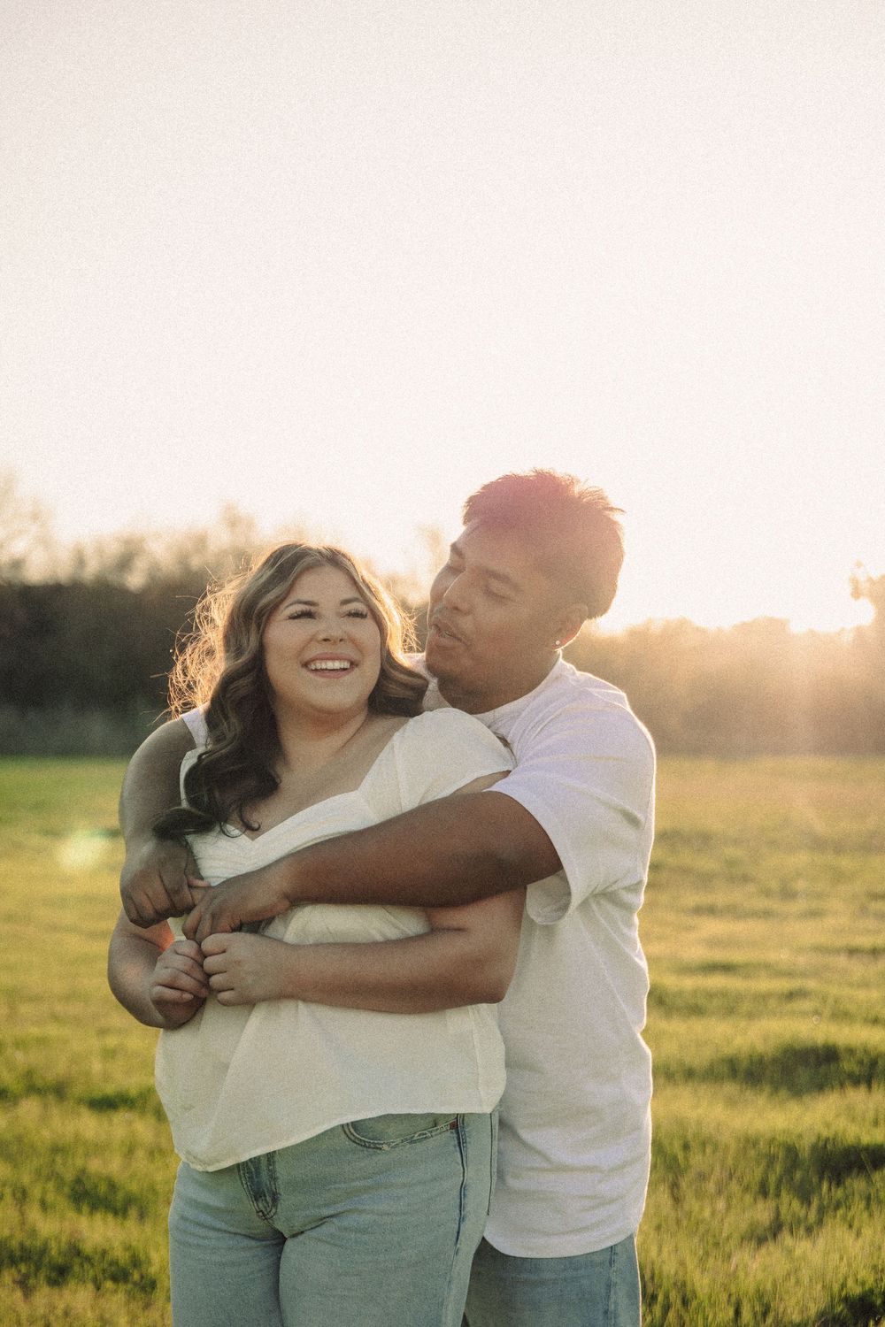 A couple embraces and laughs together in a sunlit grassy field during golden hour.