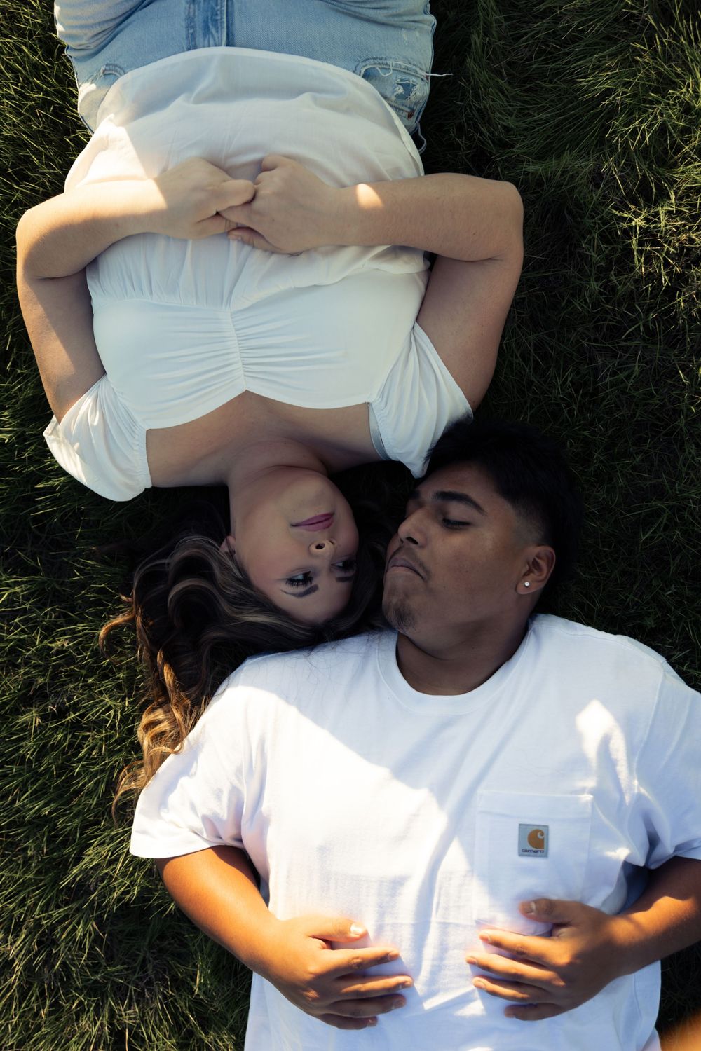 A couple lying together on green grass, wearing white clothing in a romantic outdoor setting.