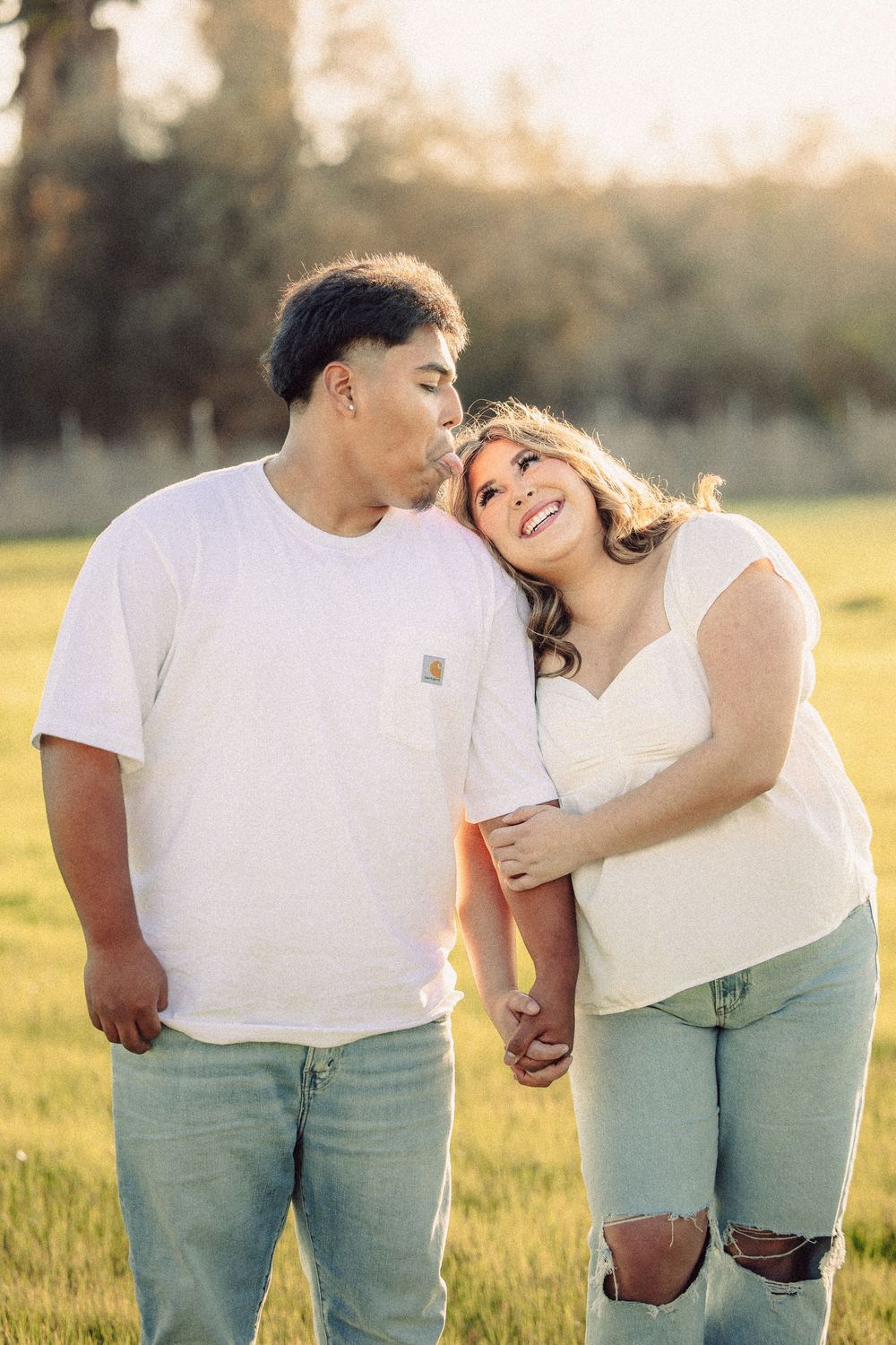 A couple in white shirts and jeans embraces in a sunlit field during golden hour.