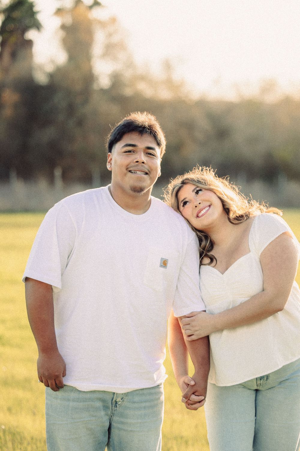 A couple in casual white tops and jeans embrace while standing in a sunlit grassy field at golden hour.