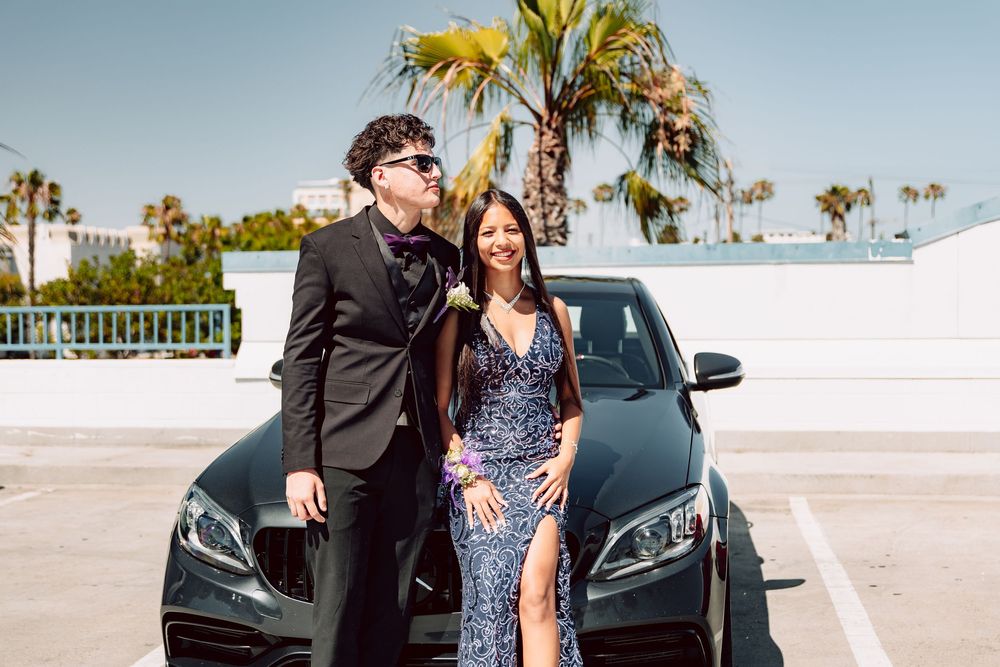 Couple in formal prom attire poses in front of black Mercedes in sunny parking lot.