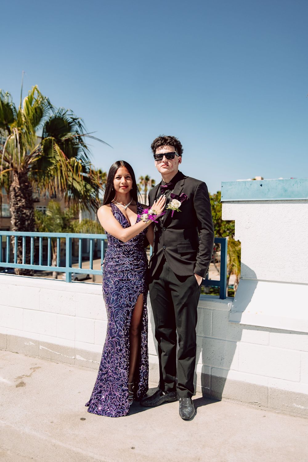 A couple dressed in prom attire, woman in purple sequin gown and man in black suit, posing outdoors with palm trees.