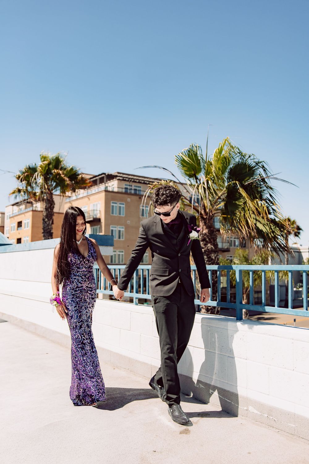 A prom couple in elegant formalwear holding hands and walking along an outdoor terrace with palm trees in background.