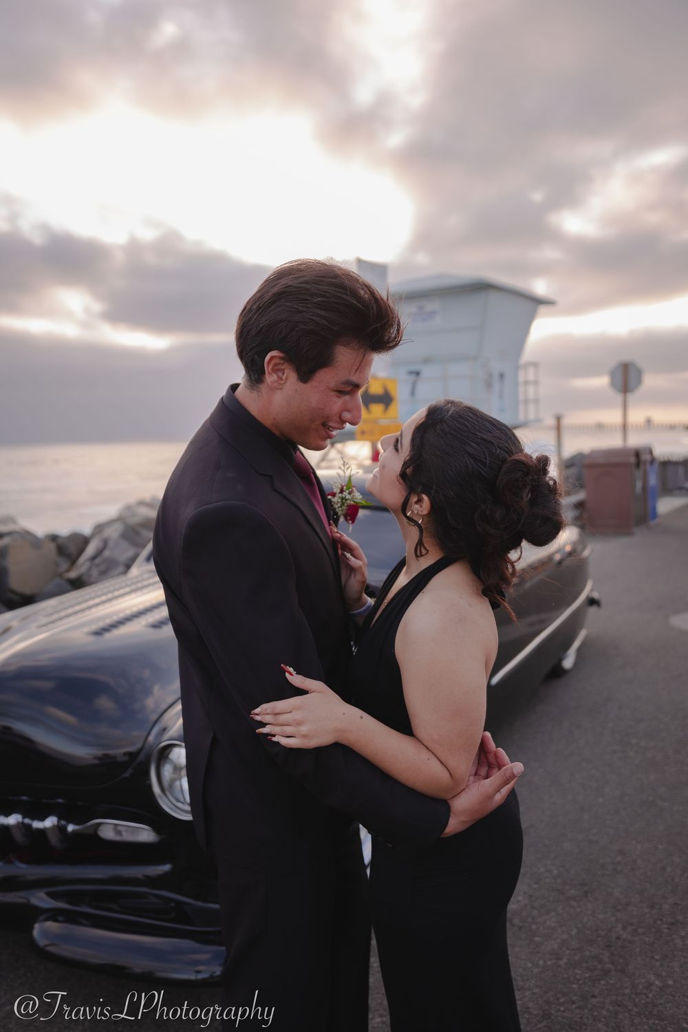 Romantic couple embracing near vintage cars at dusk with cloudy sky backdrop.