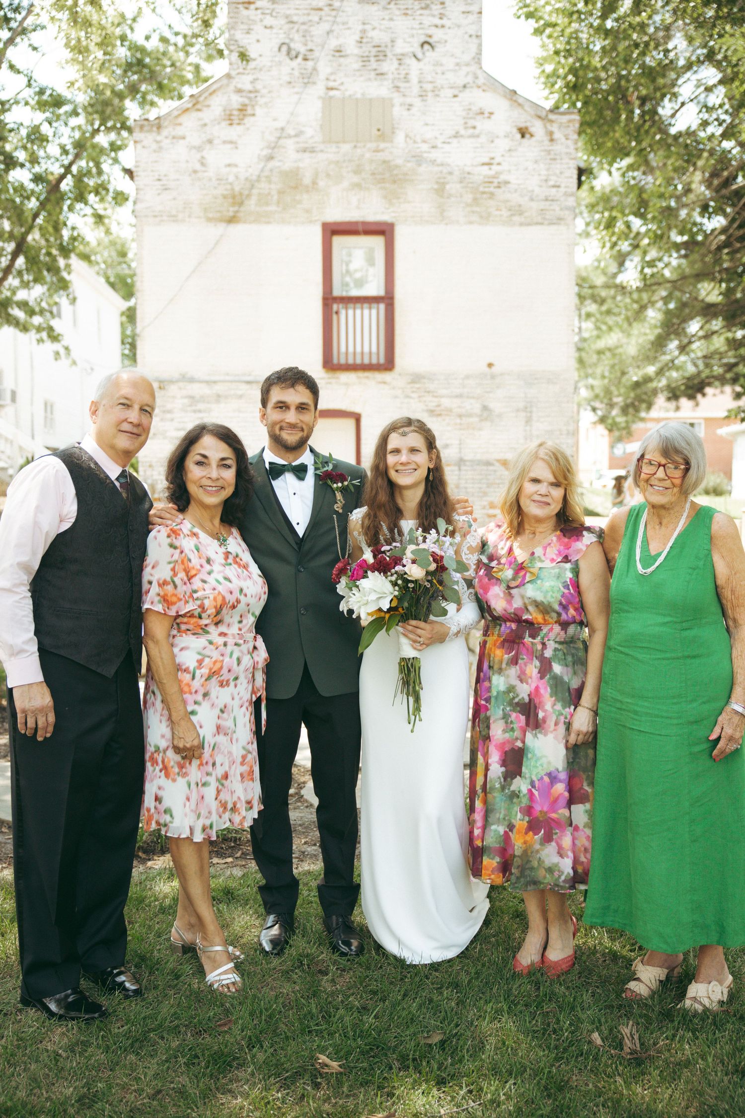 Wedding party portrait outside white stone church with floral arrangements and formal attire.