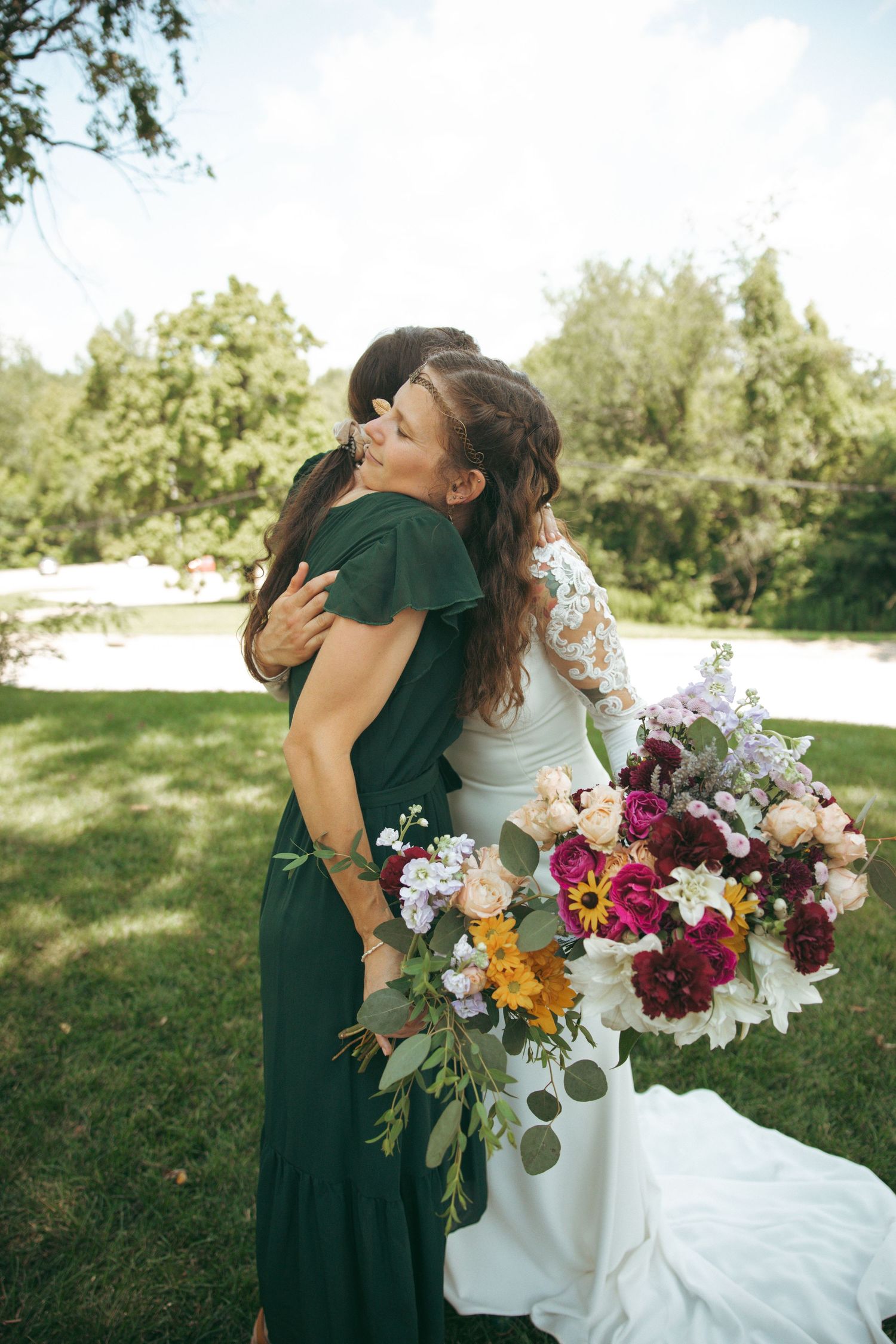 Bride in white gown embraces bridesmaid in emerald dress while holding vibrant floral bouquet outdoors.