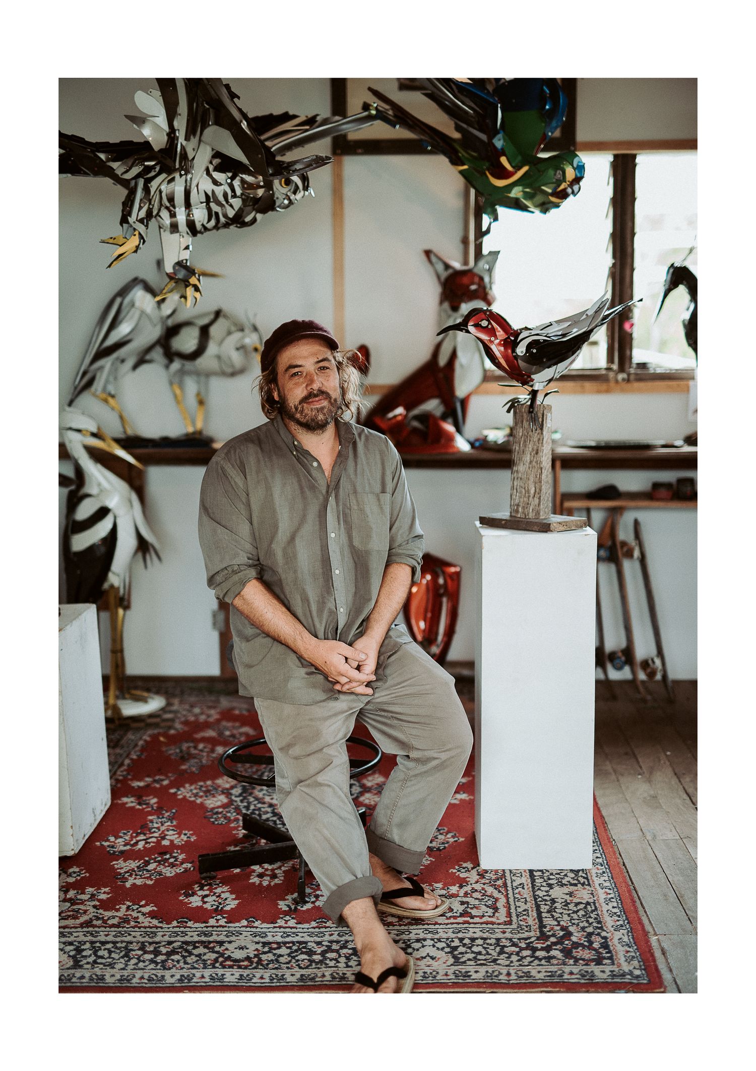 An artist sits casually in their studio surrounded by sculptures and artwork.