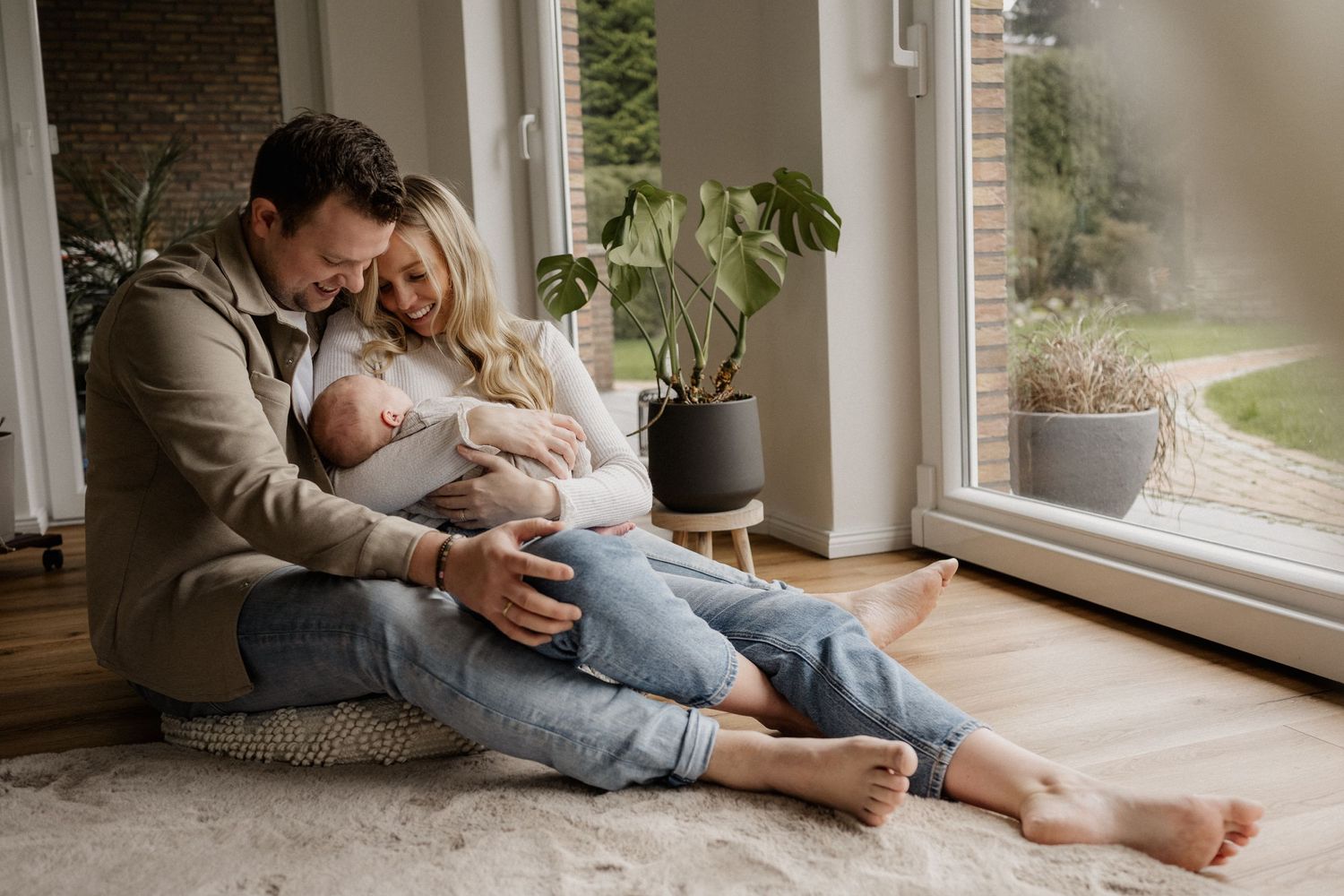 A cozy family moment with a newborn baby by large windows with natural light streaming in, featuring plants and casual comfort at home.