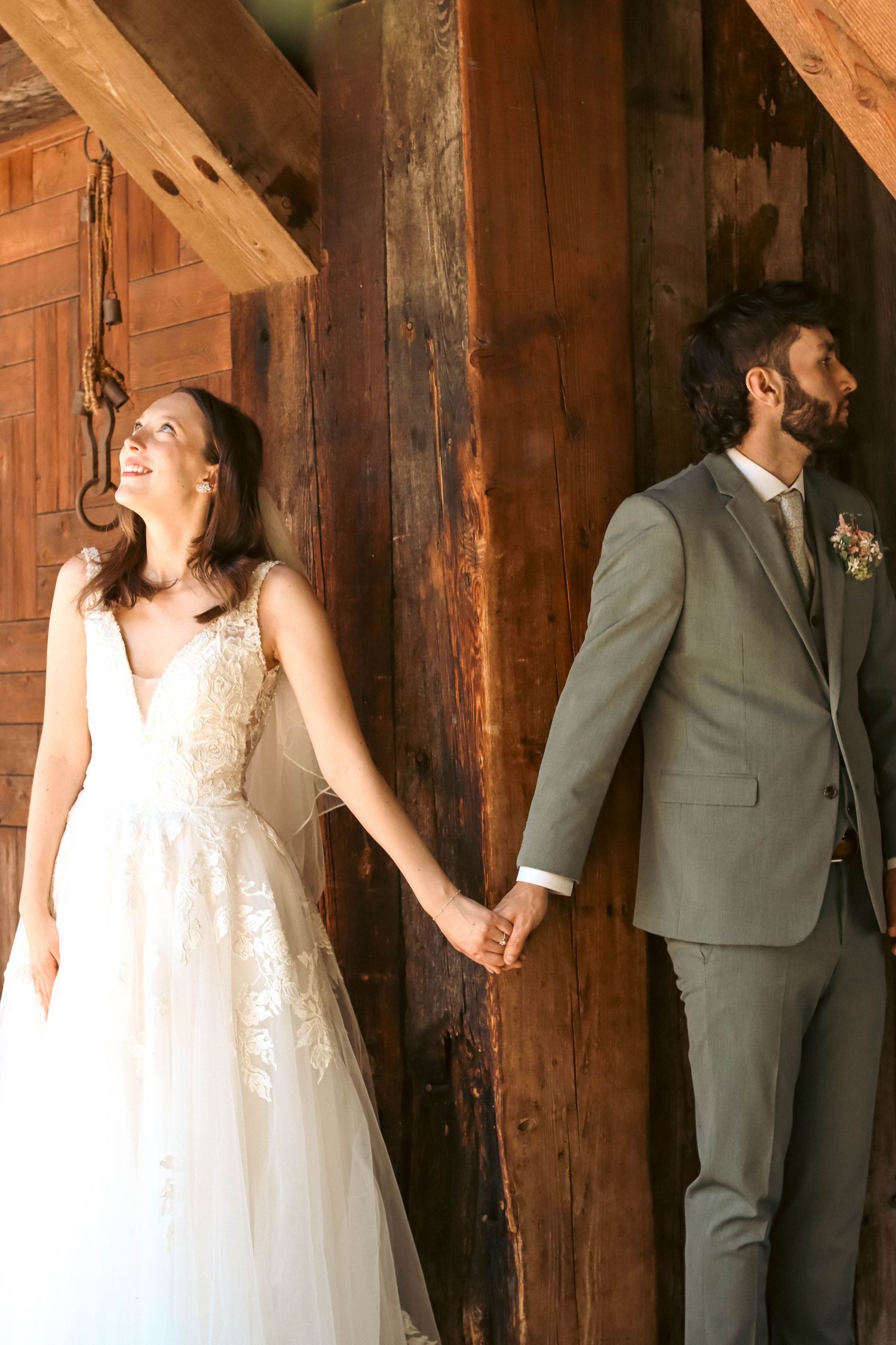 Bride in lace gown and groom in gray suit hold hands in rustic barn.