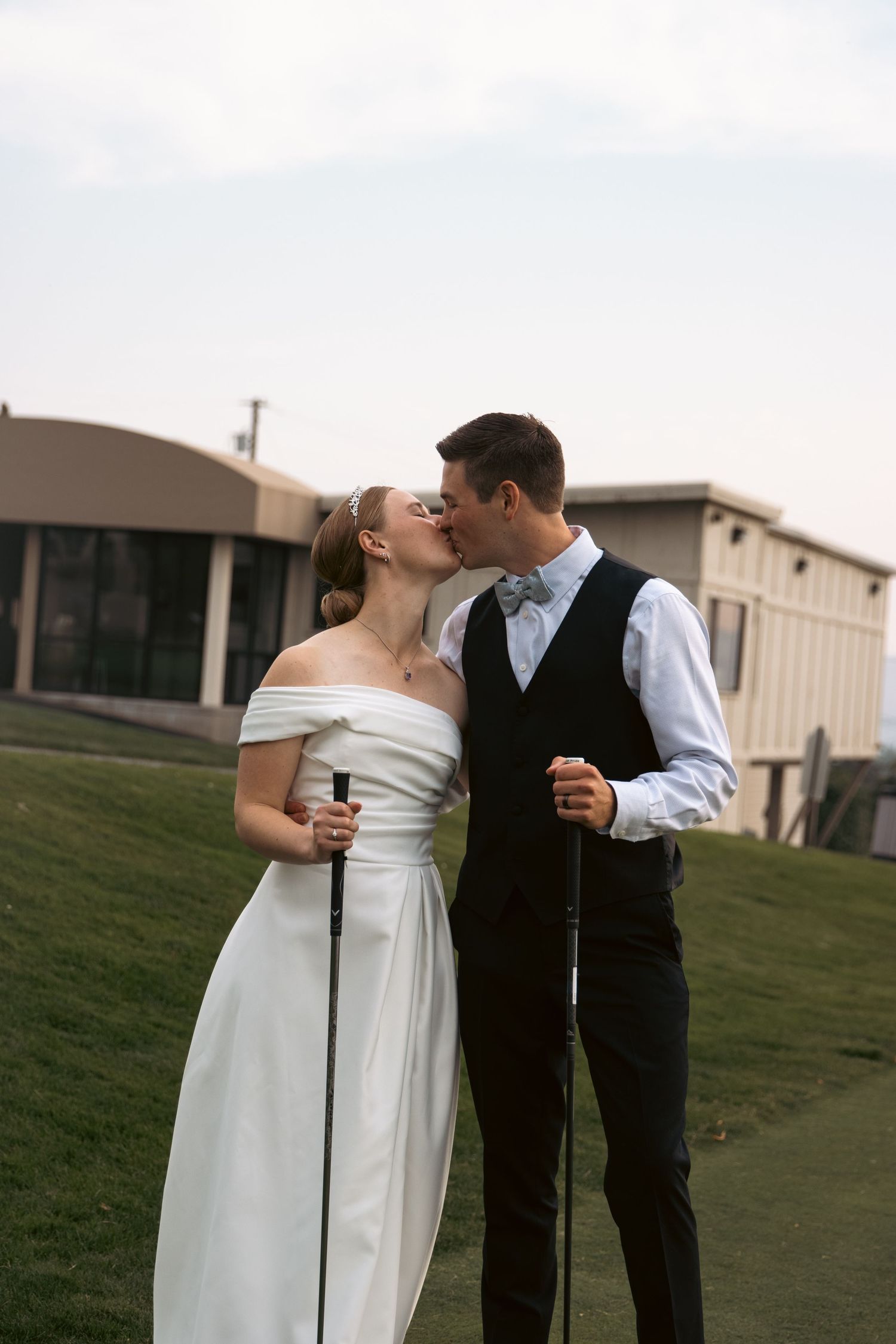 Bride and groom share a kiss on a golf course holding golf clubs on their wedding day.