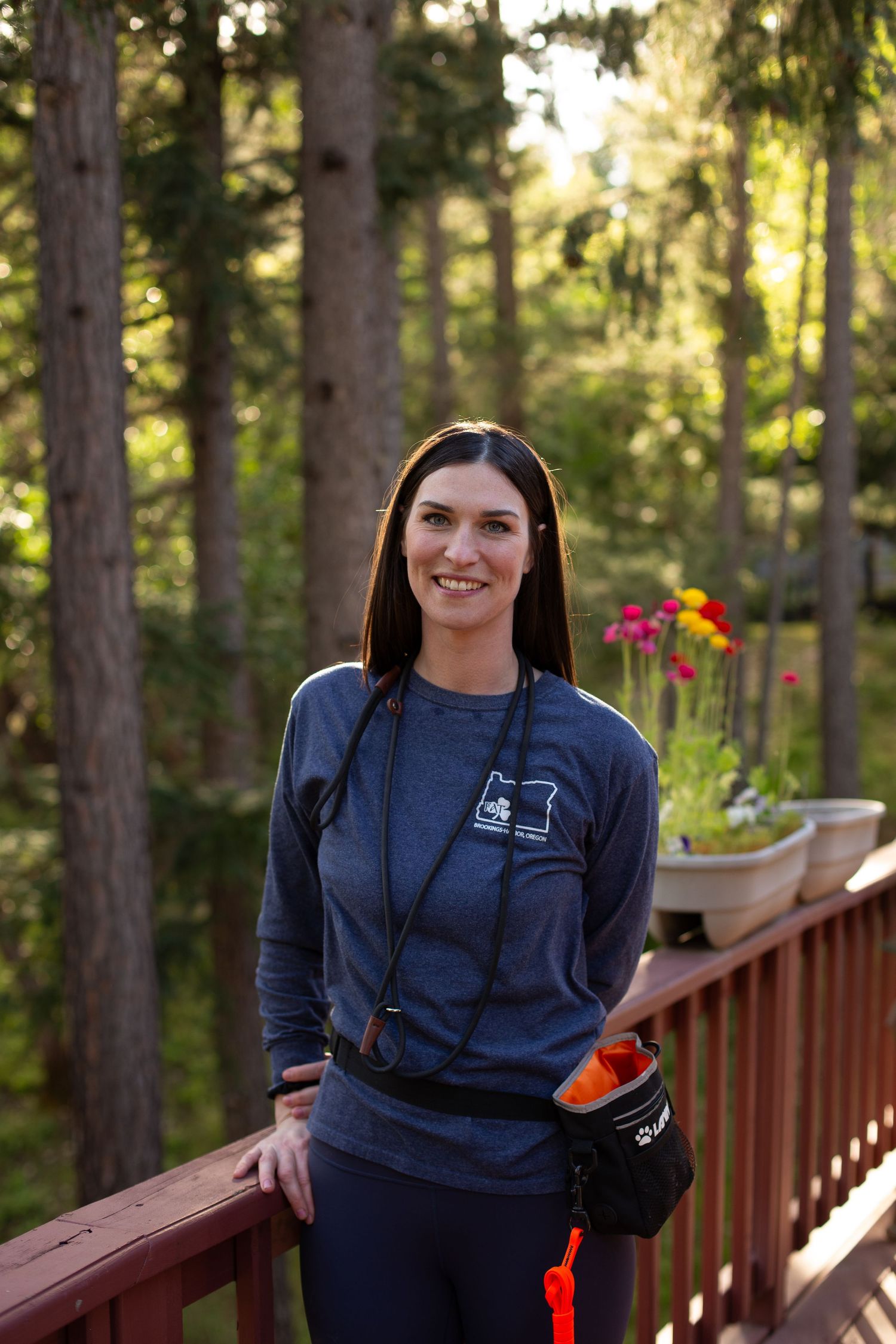 A dog trainer stands on a wooden deck in casual athletic wear with evergreen trees and sunlit forest in the background.