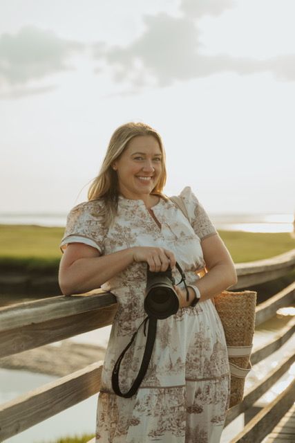 Smiling blonde female photographer holding camera on wooden bridge at golden hour.