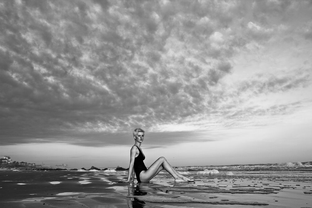 Dramatic beachside pose with cloudy sky and ocean waves in black and white photography.