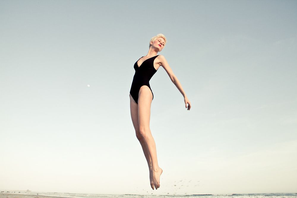 Artistic shot of figure suspended mid-jump wearing black swimsuit against bright sky.