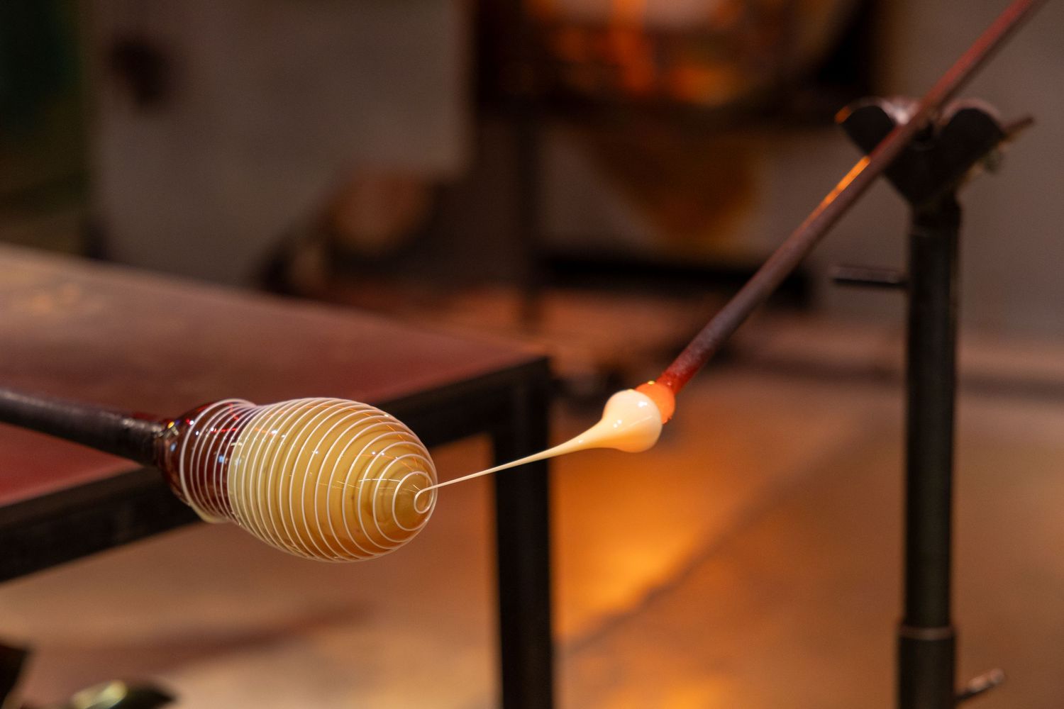 Glassblower shaping molten golden glass on metal rod in warm glowing workshop environment.