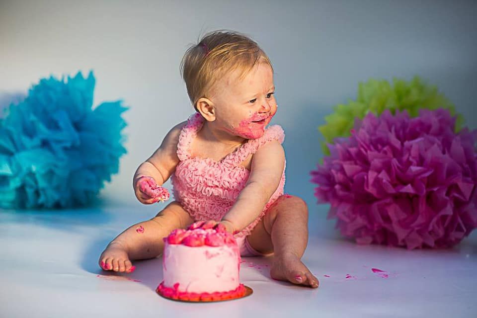 Baby in pink outfit celebrating with birthday cake surrounded by colorful tissue decorations.