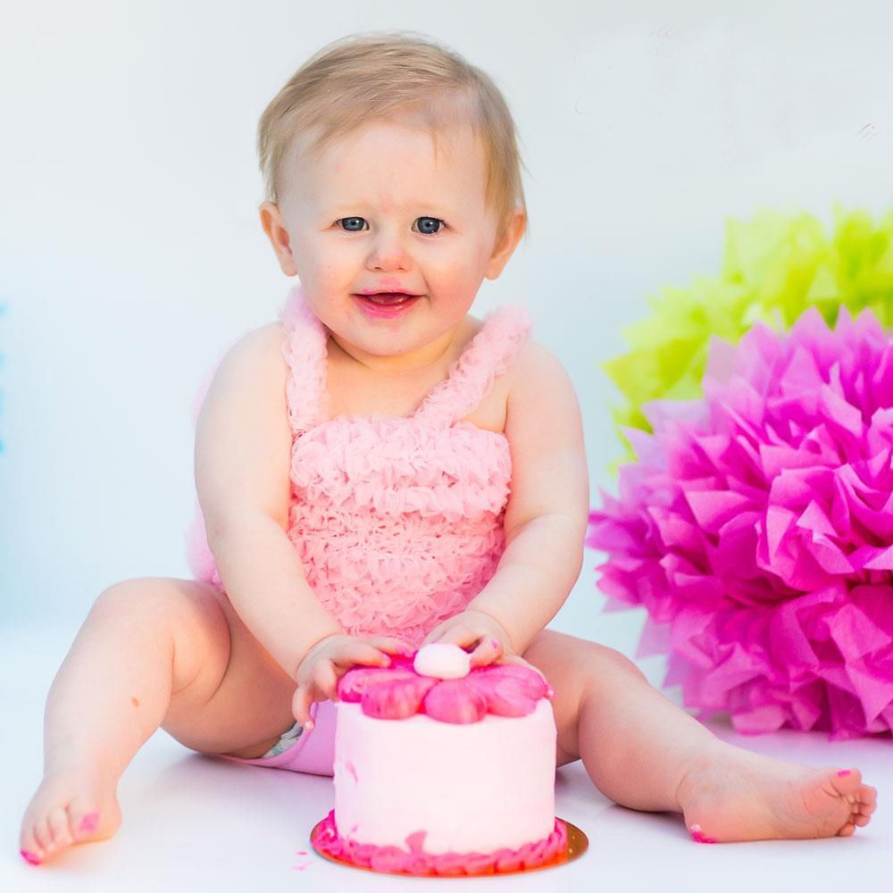 Baby in pink outfit sitting with small pink birthday cake and decorative pom poms.