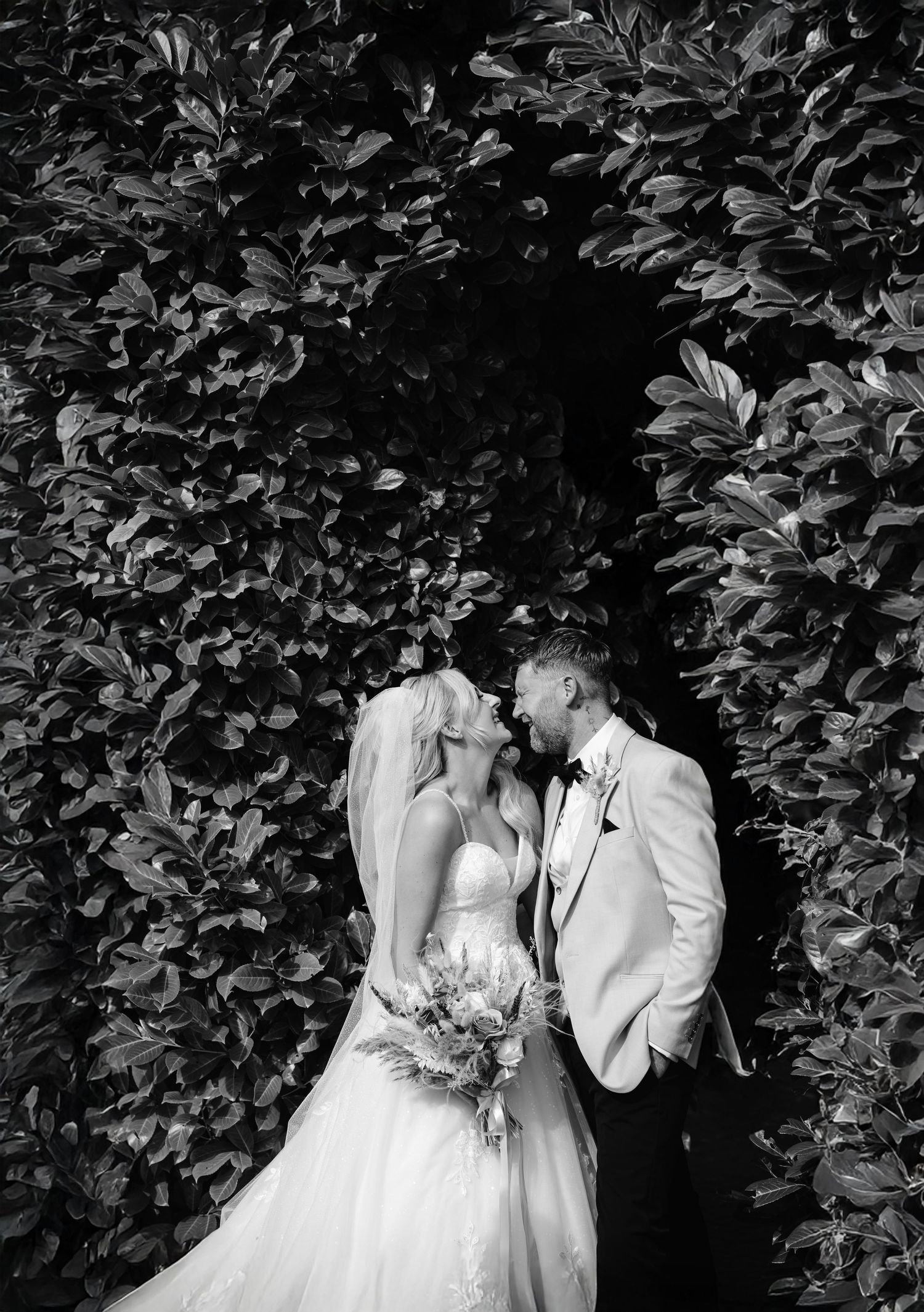 Bride and groom share a romantic moment under a lush green leafy arch in black and white.