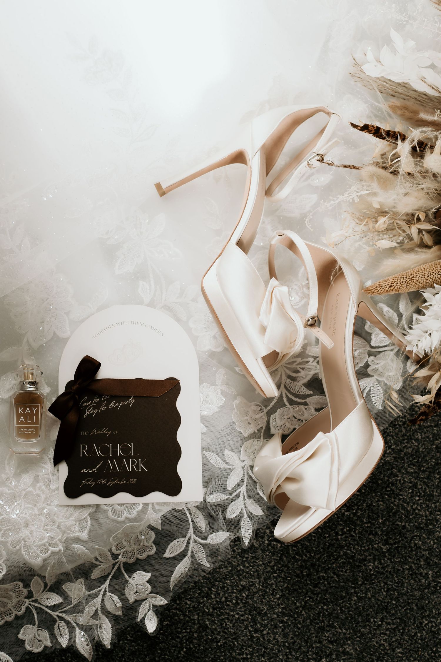 Elegant bridal details flat lay featuring ivory satin heels, wedding invitation for Rachel and Mark, and dried pampas grass.