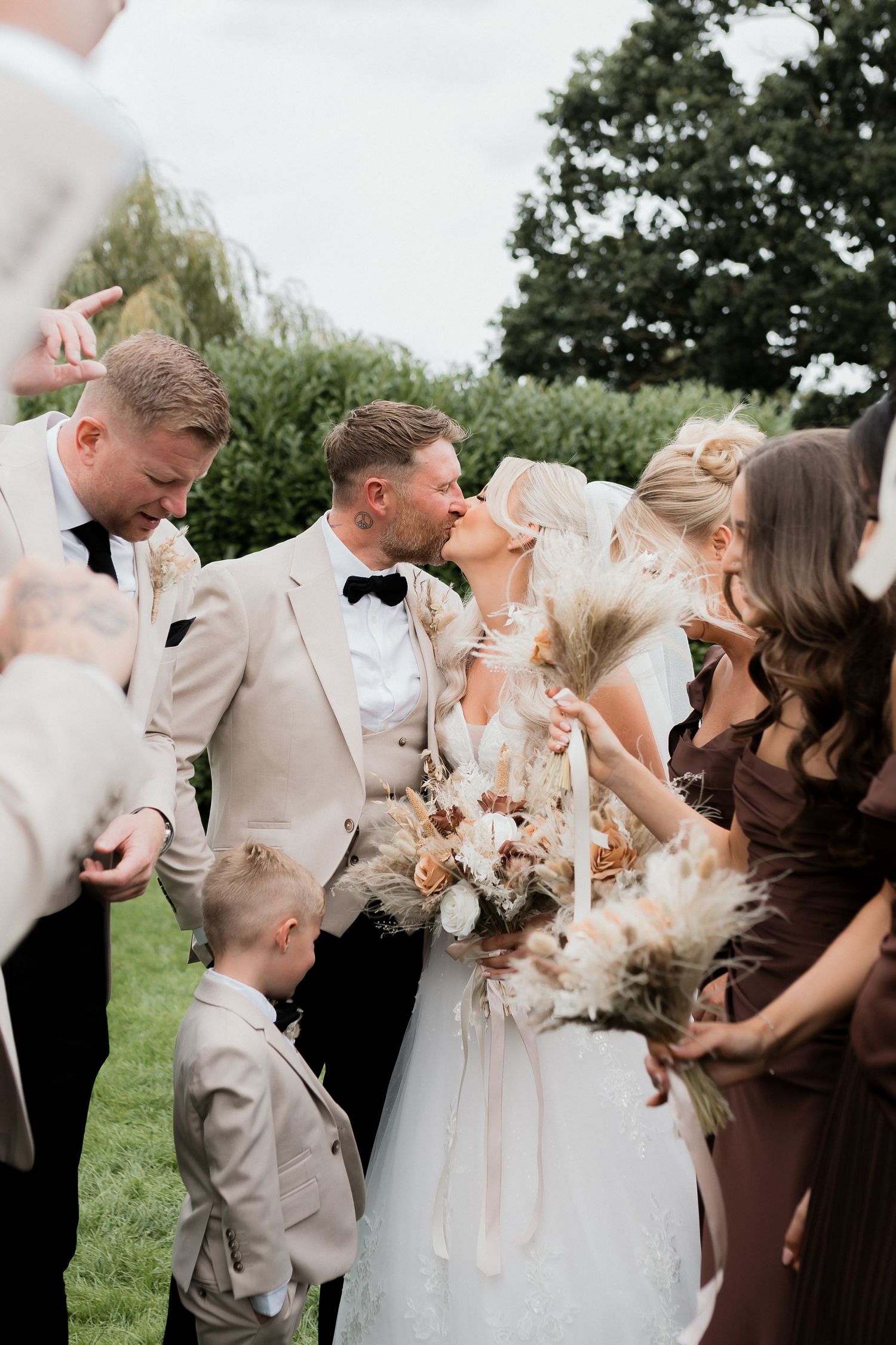 Wedding party group photo outdoors with bride and groom kissing, bridesmaids in brown dresses and groomsmen in beige suits.