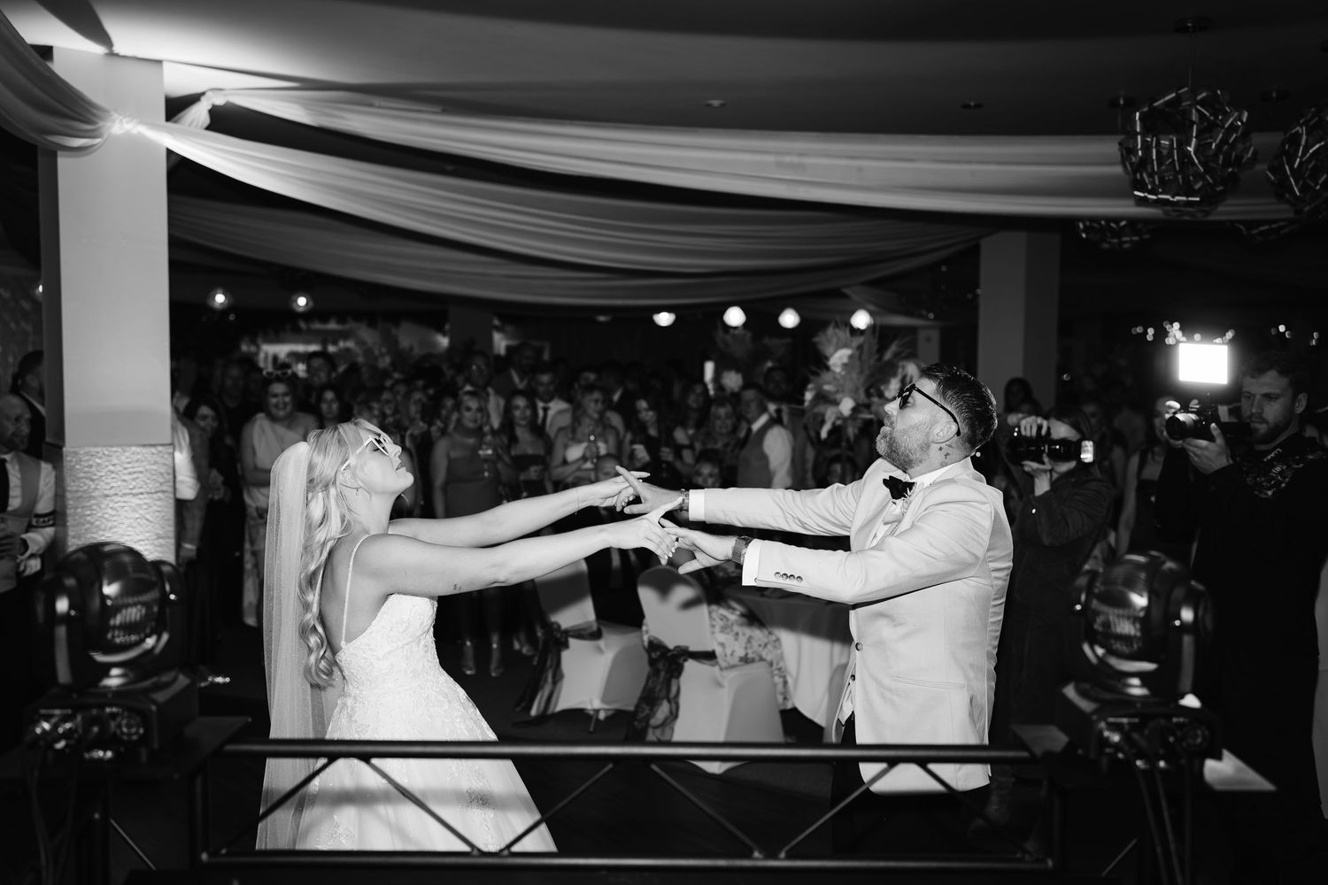 Black and white photo of bride and groom cutting wedding cake on dance floor surrounded by guests.