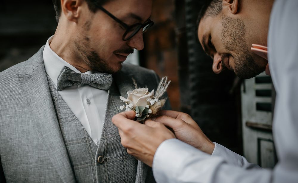 Groomsman carefully pinning a floral boutonniere onto the groom's grey suit lapel outdoors.