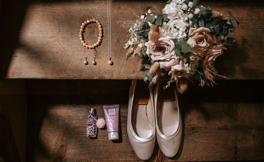 Wedding flat lay featuring bridal heels, bouquet, jewelry, and beauty products on a wooden surface.