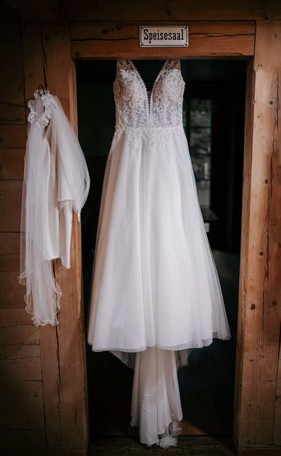 Elegant white lace wedding dress hanging in a rustic wooden doorway with a veil beside it.