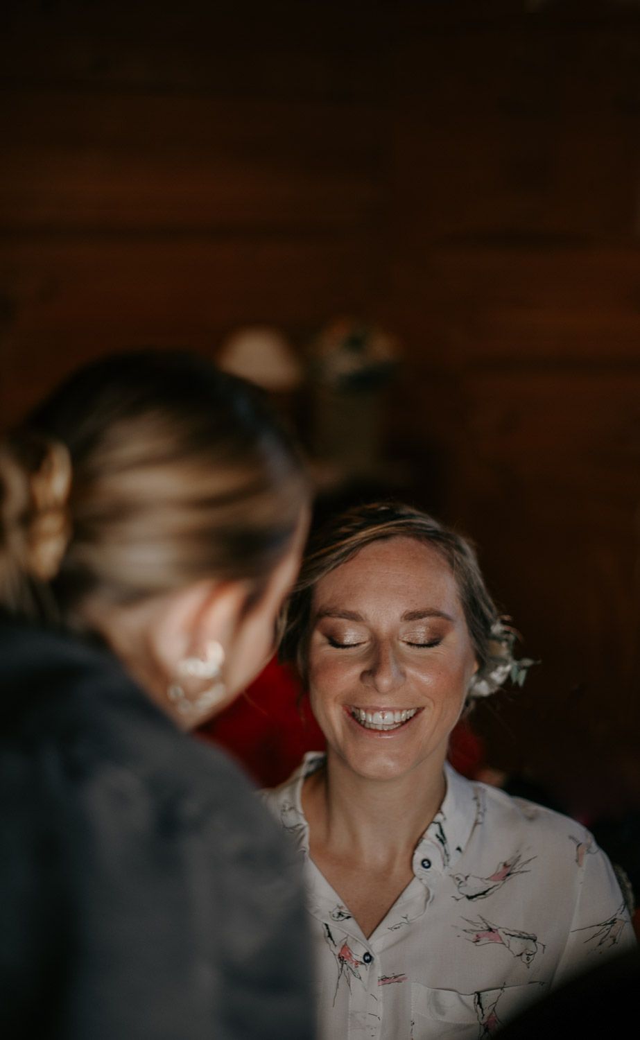 Bride smiling joyfully while having her makeup applied in a warmly lit rustic wooden interior.
