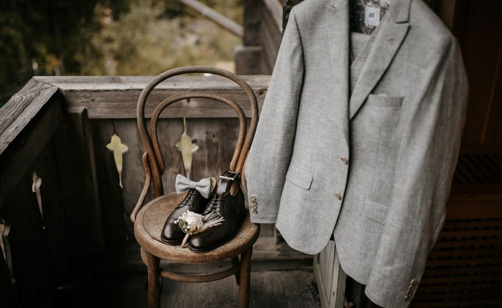 Grey groom's suit jacket hanging beside vintage shoes and a bow tie on a rustic wooden chair.