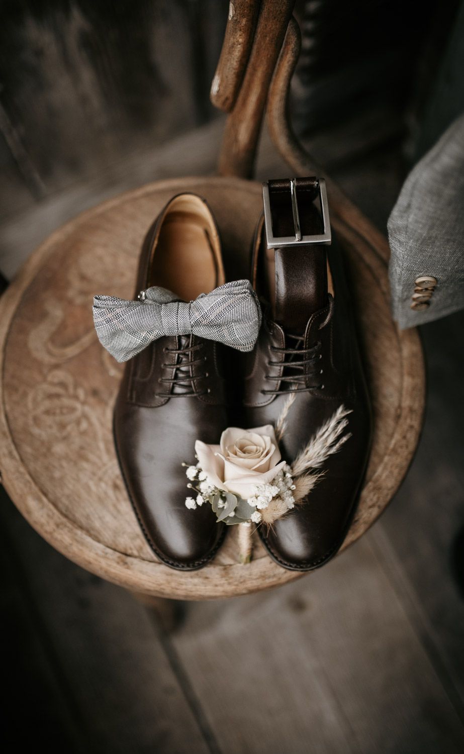 Groom's dark Oxford shoes styled with a bow tie, boutonniere, and belt on a wooden stool.