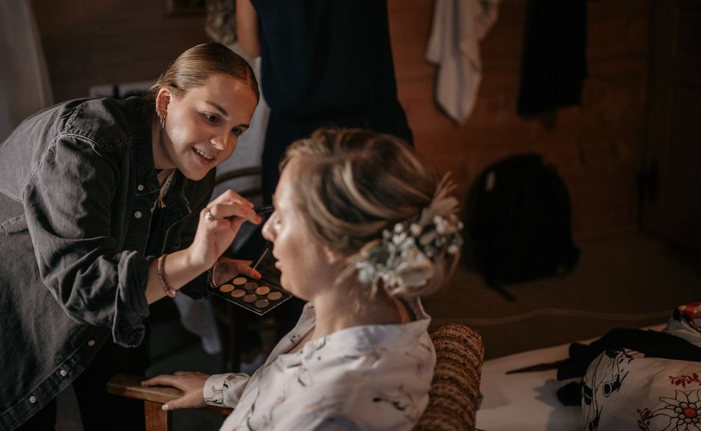 Professional makeup artist applying eyeshadow to a bride wearing a floral hair accessory during wedding preparations.