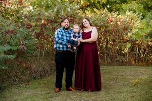 A couple poses together in a wooded outdoor setting with autumn foliage in the background.