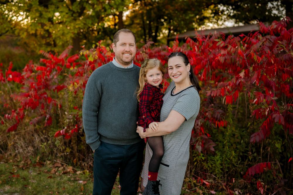 A family poses together in front of vibrant red autumn foliage during a fall photo session at sunset.