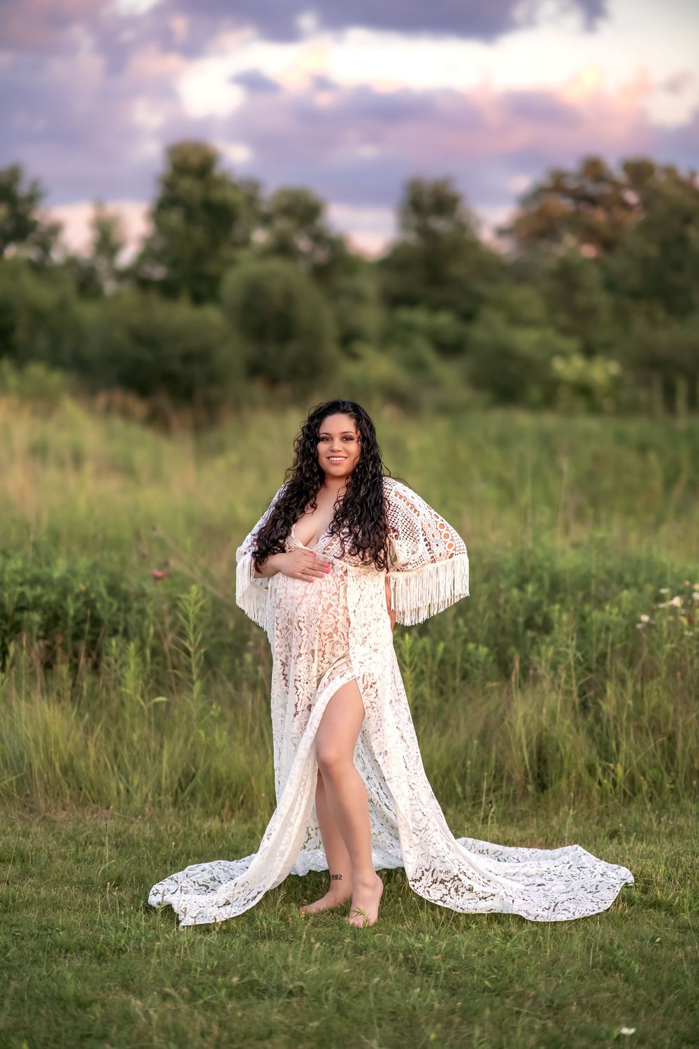 A figure in a flowing white lace dress poses gracefully in a green meadow at sunset against a backdrop of trees.