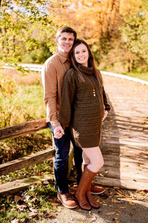 A couple poses in fall attire on a wooden walkway surrounded by autumn foliage.