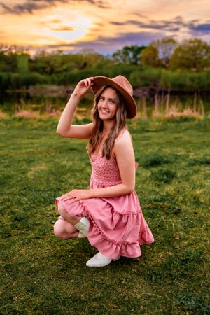 A woman in a pink dress and hat poses in a field during golden hour sunset.