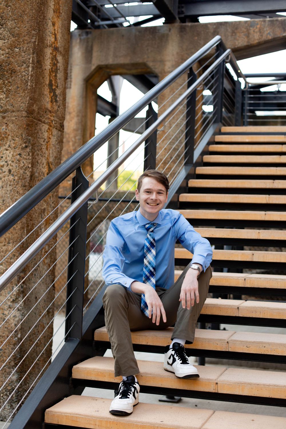 A person in a blue shirt and tie poses casually on modern wooden stairs.