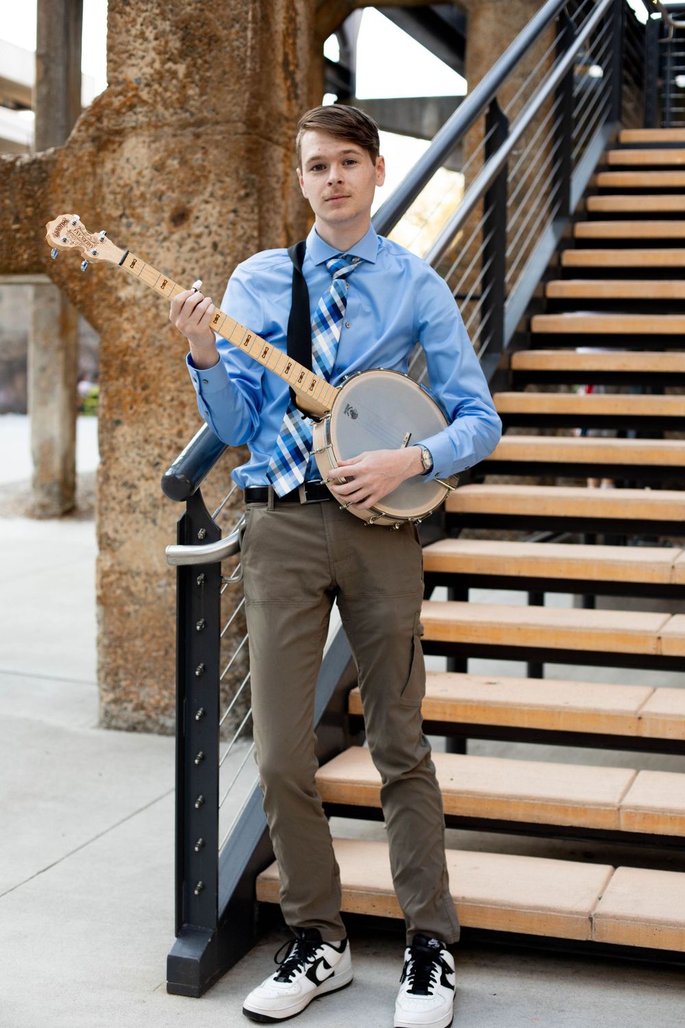 A musician poses with a banjo on industrial-style wooden stairs.