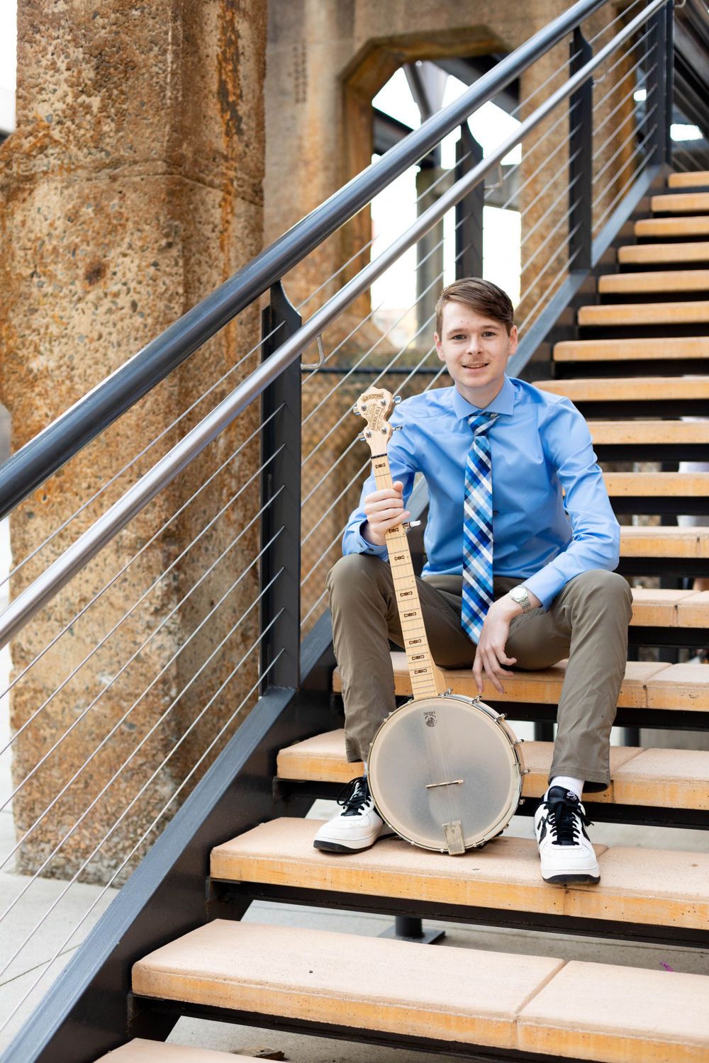 A performer sits on wooden steps holding a banjo against an industrial backdrop.