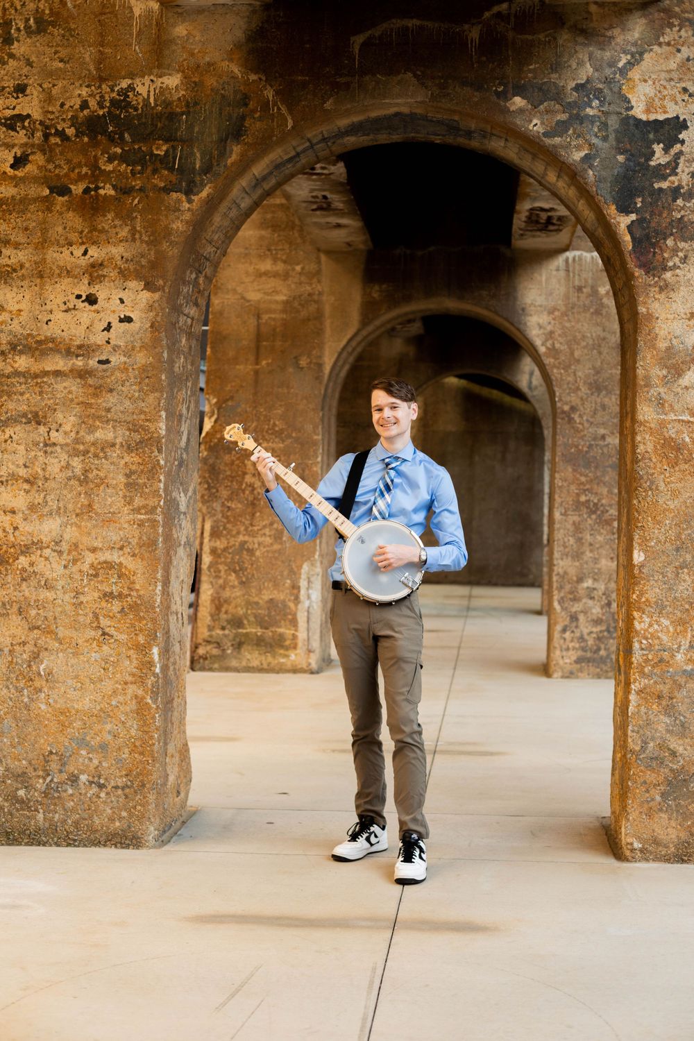 A musician poses with a banjo under historic stone archways.