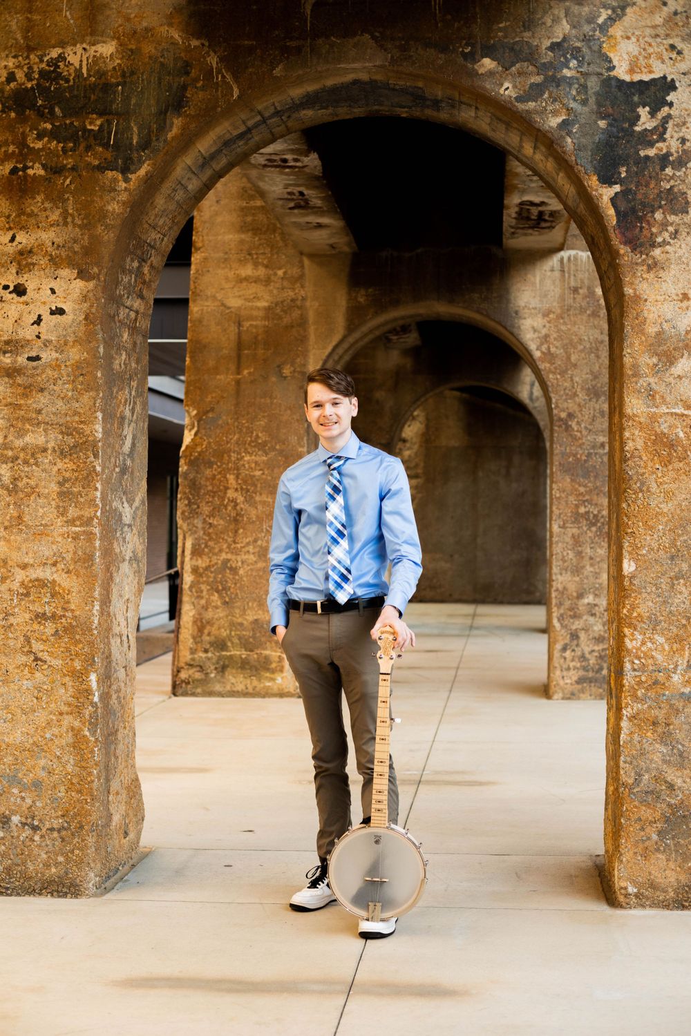A banjo player stands in an arched stone passageway wearing business casual attire.