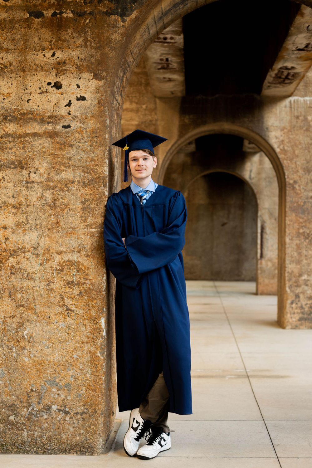 Graduate in blue cap and gown poses against stone archway in formal graduation photos.