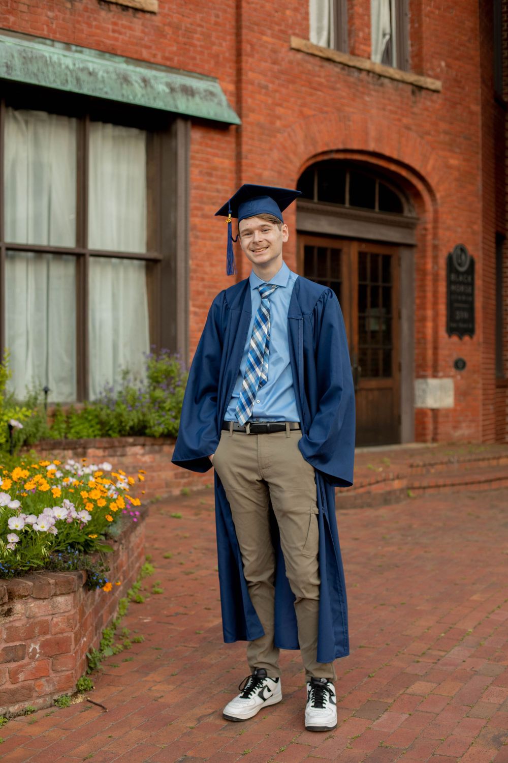 Graduate in blue regalia stands on brick pathway with flowers in background.