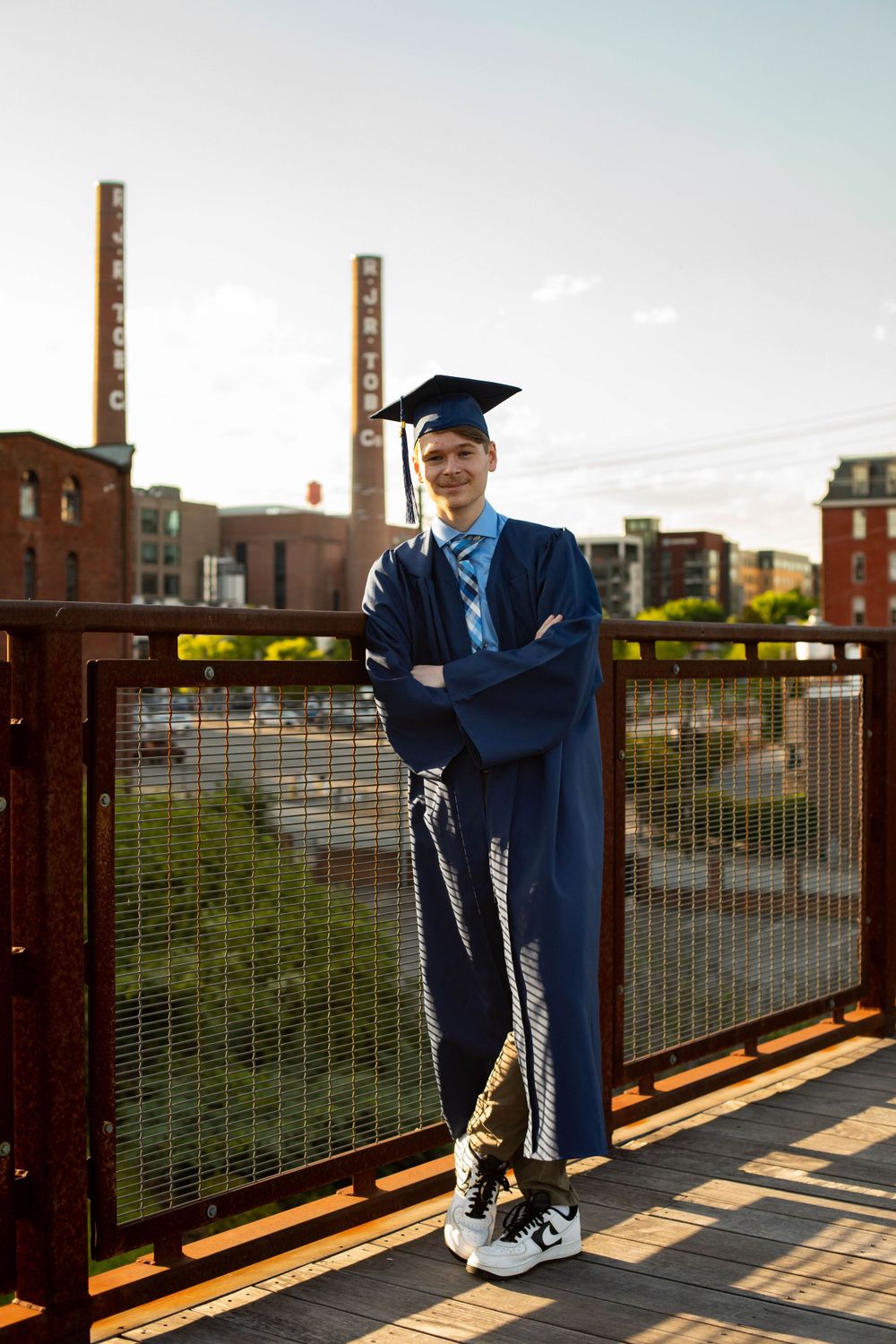 Graduate in blue academic regalia poses on industrial bridge during sunset.
