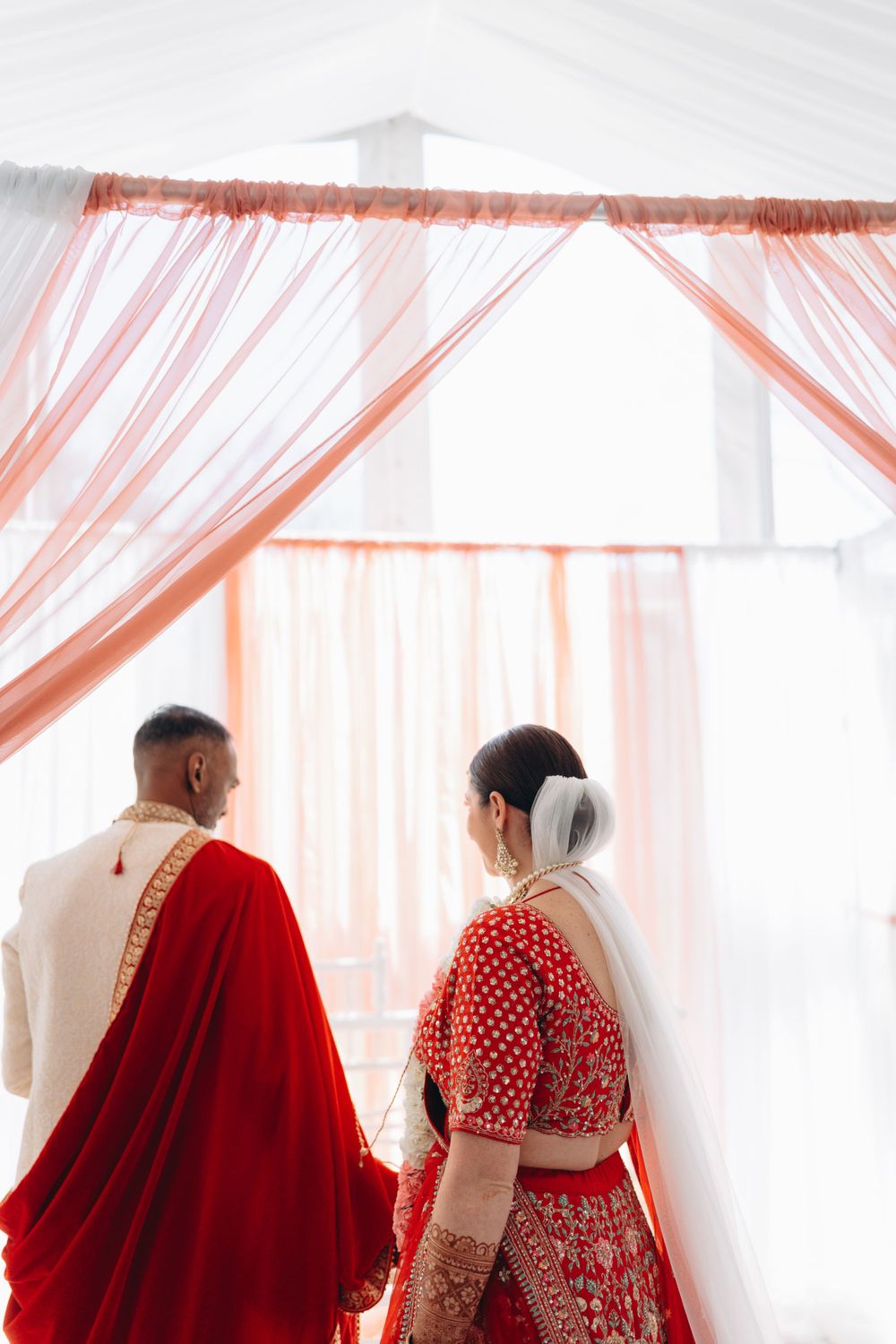 A couple in traditional South Asian wedding attire standing by sheer peach curtains in an elegant setting.